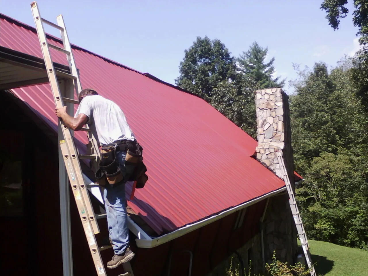 Roofer on a ladder beside a red metal roof and stone chimney — Soffit & Fascia Repair by Roofing Brothers in Suisun City